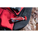 A Leatherman Skeletool RX Multitool in red is clipped to a red and black fabric—likely a backpack—with a zipper nearby, set against a rocky background.
