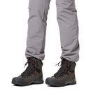 A person in light gray pants and dark gray Columbia Mens GeoTerra OutDry Leather Hiking Boots with black soles and brown laces stands against a white background. Only the lower legs and feet are visible. Vibram outsoles deliver superior grip.