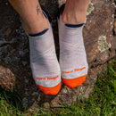 A person with a tattoo on their ankle wears Darn Tough Mens Run No Show Tab Cushioned Ultra-Lightweight Running Socks in white and orange while standing on a rock outdoors, surrounded by grass.