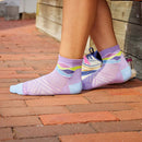 Wearing Darn Tough Womens Run Quarter Ultra-Lightweight Running Socks in a colorful pattern with sneakers, standing on a brick walkway by a wooden wall. These socks are moisture-wicking to keep feet dry and comfortable all day.