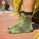 A person stands on a brick sidewalk wearing Darn Tough Mens Vert Micro Crew Ultra-Lightweight Socks featuring green Merino wool with black and orange cicada designs. Greenery and a street with a stop sign are visible in the background.