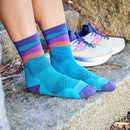 A person wearing Darn Tough Womens Mirnavated Micro Crew Ultra-Lightweight Running Socks with a vibrant blue, purple, and orange pattern sits on a rock, white running shoes set nearby on the ground.