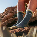 Close-up of a person’s lower legs wearing Darn Tough Mens Hiker Micro Crew Midweight Hiking Socks with a mountain design in green and yellow, standing on rocky terrain under sunlight.