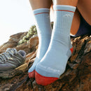 A woman wearing Darn Tough Womens Hiker Micro Crew Midweight Hiking Socks in light blue with red toes and heels sits on sunlit rocks outdoors, gray sneakers nearby. The midweight design ensures a blister-free hiking experience.