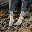 A person sits on rocks outdoors, wearing Darn Tough Mens Mountaineering Micro Crew Heavyweight Hiking Socks in a light color with rolled-up pants. Behind them, mountains rise above tree-lined and grassy slopes.