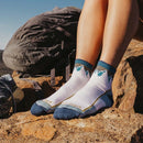 A person wearing Darn Tough Womens Hiker Quarter Midweight Hiking Socks in white and blue sits on rocks beside a gray backpack, sunlight illuminating their legs and outdoor gear in the background.