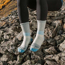 A person sits on rocky terrain outdoors amid autumn foliage, wearing black leggings and Darn Tough Womens Mountaineering Micro Crew Heavyweight Hiking Socks in a gray-blue hue.