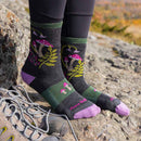 A person wearing black leggings and Darn Tough black socks with pink and green mushroom designs stands on a rocky surface outdoors; a pair of Womens Mellow Mushrooms Micro Crew Lightweight Hiking Socks, crafted from soft Merino wool by Darn Tough, are placed beside their feet along with sturdy hiking shoes.