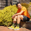 A man wearing Darn Tough Mens Vert Micro Crew Ultra-Lightweight Socks sits outdoors on a stone bench, smiling, with greenery and a wooden building behind him. He pairs the socks with an orange T-shirt, black shorts, and yellow sneakers.