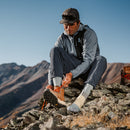A person sits on rocky mountain terrain under clear skies, wearing outdoor gear and sunglasses, as they tie the laces of their tan hiking boots; their feet stay comfortably protected by Darn Tough Mens Mountaineering Micro Crew Heavyweight Hiking Socks.