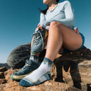 Wearing Darn Tough Womens Hiker Quarter Midweight Hiking Socks, a woman in athletic wear sits on mountain rocks adjusting her socks. She sports a light blue long-sleeve shirt, shorts, and has a backpack beside her under a clear sky.