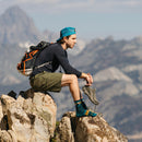 A man in a blue cap, dark shirt, and shorts sits on a mountain outcrop, holding his hiking shoes with Darn Tough Mens Sunset Ridge Micro Crew Lightweight Hiking Socks visible, gazing thoughtfully at the scenic landscape.