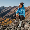 Perched on rocky terrain atop a mountain, a woman in a blue jacket smiles as she switches into Darn Tough Womens Mountaineering Micro Crew Heavyweight Hiking Socks, savoring their warmth and comfort after a long adventure. In the background, scenic mountains and vibrant fall foliage stretch beneath the clear sky, while she holds her hiking boots in hand.