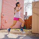 A woman in a white tank top and pink shorts jogs on a sunny day past a peach-colored wooden house and fence, wearing Darn Tough Womens Mirnavated Micro Crew Ultra-Lightweight Running Socks, with leafless branches in the background.