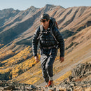 A man wearing outdoor gear, sunglasses, and Darn Tough Mens Mountaineering Micro Crew Heavyweight Hiking Socks treks along a rocky mountain trail, surrounded by autumn-hued trees and rugged, sunlit peaks in the background.