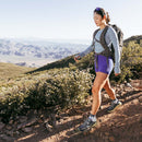 A woman in athletic wear and Darn Tough Womens Hiker Quarter Midweight Hiking Socks hikes a rocky trail lined with green shrubs, mountains, and clear sky in the background.