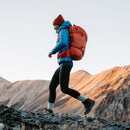 A person wearing a blue jacket, orange backpack, red beanie, and Darn Tough Womens Mountaineering Micro Crew Heavyweight Hiking Socks hikes up a rocky slope with mountains in the background under a clear sky.