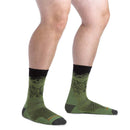 A person wearing Darn Tough Mens Vert Micro Crew Ultra-Lightweight Socks stands on a white background. The green and black socks feature a mountain design with dark toes, heels, and cuffs. Only the lower legs and feet are visible.