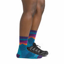 A person wearing Darn Tough Womens Mirnavated Micro Crew Ultra-Lightweight Running Socks, featuring colorful patterns in blue, purple, orange, and teal with black athletic shoes, stands on a white background.
