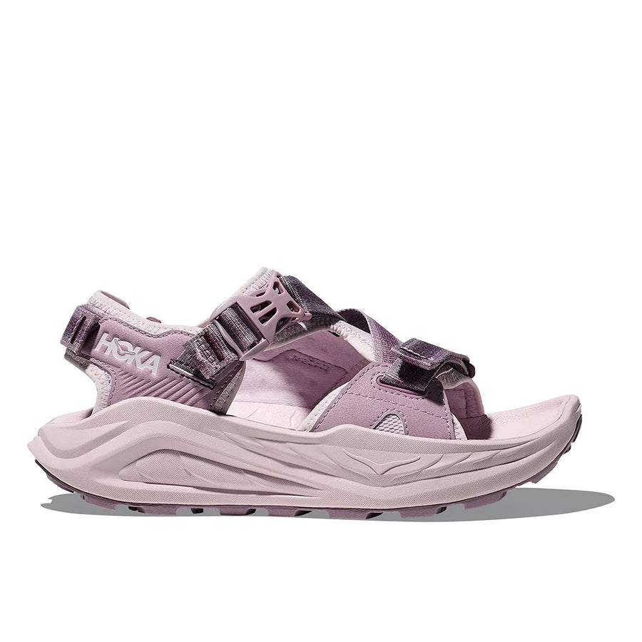 The Womens Infini Hike TC Trail Sandal by Hoka is a light purple sandal inspired by Hoka running shoes, featuring adjustable straps, an open toe, thick cushioned sole, and rugged tread, displayed in a side view on a white background.