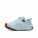 Light blue Hoka Womens Transport 2 athletic sneakers with a thick, textured brown sole. These city-to-trail shoes feature a low-cut design, visible stitching, and tags labeled “Gore-Tex” and “Vibram.” Designed for seamless transitions from urban streets to rugged paths. Background is plain white.