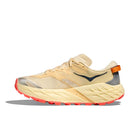 The Hoka Womens Speedgoat 7 Trail Running Shoe features beige and yellow colors, a thick textured sole, orange accents like a heel pull tab, and red tread on the outsole.