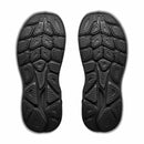 A pair of black shoe soles featuring textured, geometric tread patterns are shown from the bottom against a white background—these are the distinctive outsoles of the Hoka Womens Clifton 11 Wide Running Shoe, designed to deliver superior comfort for women runners.