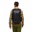 A person wearing a brown plaid shirt and green pants is shown from behind, carrying a large black Cotopaxi Allpa 35L Travel Pack—crafted from recycled fabrics—with a water bottle tucked into the side pocket.