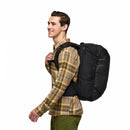 A person in a plaid shirt and green pants smiles while carrying the Cotopaxi Allpa 35L Travel Pack—crafted from recycled fabrics—over one shoulder, walking to the left against a plain white background.