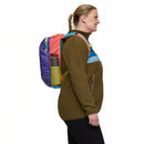 A person in a brown fleece jacket and black pants stands in profile, wearing a colorful Cotopaxi Allpa Mini 20L Travel Pack - Del Día with a green water bottle in the side pocket. The background is plain white.