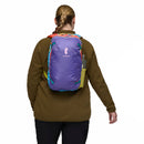 A person with light brown hair tied in a ponytail, dressed in a brown jacket, is seen from behind carrying the Cotopaxi Allpa Mini 20L Travel Pack - Del Día and holding a beverage in their left hand.