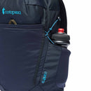 Close-up of a dark blue Cotopaxi Elqui 18L Backpack featuring a side pocket holding a black water bottle with a red cap. The Cotopaxi logo and blue bungee cord are visible on this recycled nylon backpack.