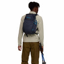 A person stands facing away against a white background, wearing a gray Cotopaxi Elqui 18L Backpack and a beige long-sleeve shirt, holding blue trekking poles in their right hand.