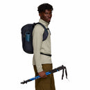 A person wearing outdoor gear with a Cotopaxi Elqui 18L Backpack and holding blue trekking poles, standing sideways against a white background.