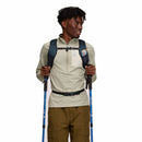 A person with short curly hair, dressed in a light long-sleeve shirt and brown pants, stands against a plain white background holding blue trekking poles and wearing the Cotopaxi Elqui 18L Backpack in black.