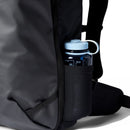 A close-up of the Cotopaxi Allpa 35L Travel Pack highlights a blue water bottle tucked into its side mesh pocket. The water bottle is designed with a screw cap, loop handle, and clear measurement markings. Meanwhile, the backpack’s sleek, durable exterior—made from recycled materials—underscores Cotopaxi’s commitment to both functionality and sustainability.