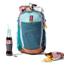 A Cotopaxi Elqui 18L Backpack - Del Día, filled with snacks, a chocolate bar, and water bottles, sits next to a glass bottle of Coca-Cola with a straw and a bag of potato chips spilled out on the white surface.