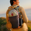 A person in a beige jacket stands outside at sunset, facing away, wearing the Cotopaxi Elqui 18L Backpack - Del Día with a water bottle and a round collapsible dish attached, overlooking a grassy area by the sea.