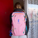 A person with long hair, wearing a black jacket and carrying a large pink Cotopaxi Allpa 28L Travel Pack - Del Día, stands facing a red door. The sustainable backpack is decorated with small pins, and the person is partially outside a building with a patterned curtain.