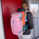 A person with a blonde braid wears a black puffy jacket and light pants, carrying the Cotopaxi Allpa 28L Travel Pack - Del Día, while standing in front of a red door.