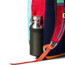 A stainless steel water bottle is in the side pocket of a colorful Cotopaxi Allpa Mini 20L Travel Pack - Del Día, featuring red, blue, orange, and mint panels against a white background.