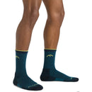 A person stands on a white background wearing Darn Tough Mens Hiker Micro Crew Midweight Hiking Socks. Only the lower legs and feet in these Merino wool hiking socks are visible. The socks feature teal color with yellow accents and a mountain design.