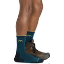 A person from the knees down wears Darn Tough Mens Hiker Micro Crew Midweight Hiking Socks—teal and navy with yellow mountain designs—standing with one foot in a brown hiking shoe, the other in just the sock.