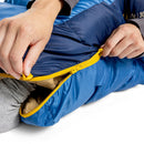 Close-up of hands unzipping the Sea to Summit Circuit Down Sleeping Bag (-1°C) in blue, featuring a yellow zipper and revealing its beige interior. The person is wearing a gray sleeve, with a white background.