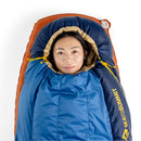 A person lies inside a Sea to Summit Circuit Down Sleeping Bag (-1°C), which is blue and tan with only their face visible. The sleeping bag is on an orange mat and features the SEA TO SUMMIT logo.
