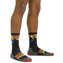 A person wearing Darn Tough Mens Sunset Ridge Micro Crew Lightweight Hiking Socks with mountain and sun designs, multicolored stripes in dark gray, orange, yellow, and green, plus contrasting toes, heels, and cuffs.