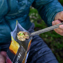 An individual wearing a blue jacket is using the MSR Titan Long Spoon to eat a meal of rice, peas, and meat from a resealable food pouch labeled ALPINE. The outdoor setting emphasizes the convenience and practicality of ultralight MSR camp cookware like the Titan Long Spoon for backcountry adventures.