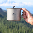 A hand holds the MSR Titan Kettle 900mL with its lid outdoors, highlighting this ultralight titanium kettle against a backdrop of blurred green trees and mountains.