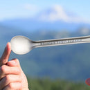 A hand holds an MSR Titan Long Spoon—the ultimate ultralight camp utensil—in front of a blurred mountain landscape and blue sky, perfectly capturing the spirit of MSR’s innovative backcountry cookware.