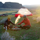 Two people sit on grass near a campfire, cooking with the MSR Titan Kettle 900mL. A red tent and two bikes are behind them, set against sunlit hills. Both wear jackets and beanies as they enjoy their outdoor camping scene, equipped with dependable backpacking cookware from MSR.